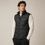A man wearing a black quilted zip-front puffer vest over a cream long-sleeve shirt and black pants, standing against a plain light beige studio background.